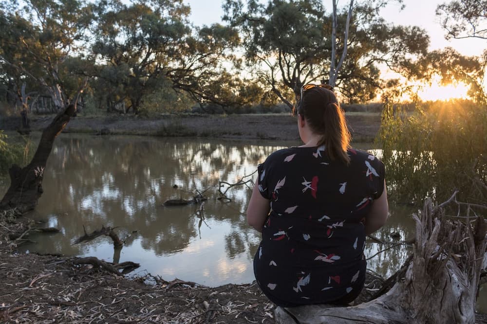 A woman sitting next to a river in quiet contemplation as the sun sets.  